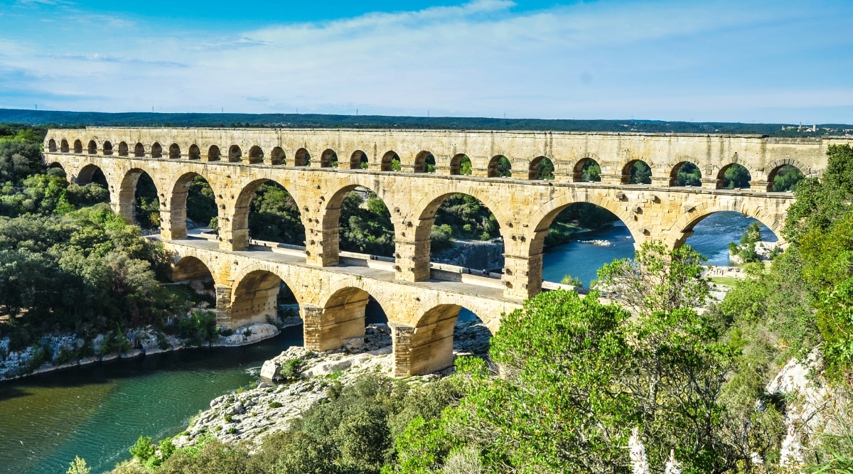 Aerial view of Pont du Gard. The Pont du Gard is a breathtaking Roman aqueduct bridge located near Nimes in the South of France. This ancient engineering spans the Gardon River with three tiers of graceful arches. The honey-colored limestone, clear blue skies, and the tranquil river below create a captivating scene.