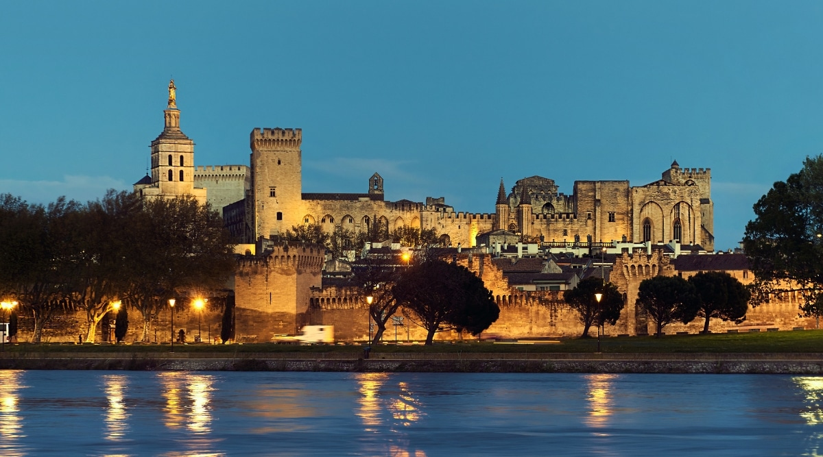 View of Remparts d'Avignon in the evening illuminated by yellow lanterns. The Remparts d'Avignon, or the city walls of Avignon, has sturdy stone walls and watchtowers. The walls are punctuated by imposing gates and battlements. In front of this medieval fortifications there is a beautiful green square with trees and a wide river.