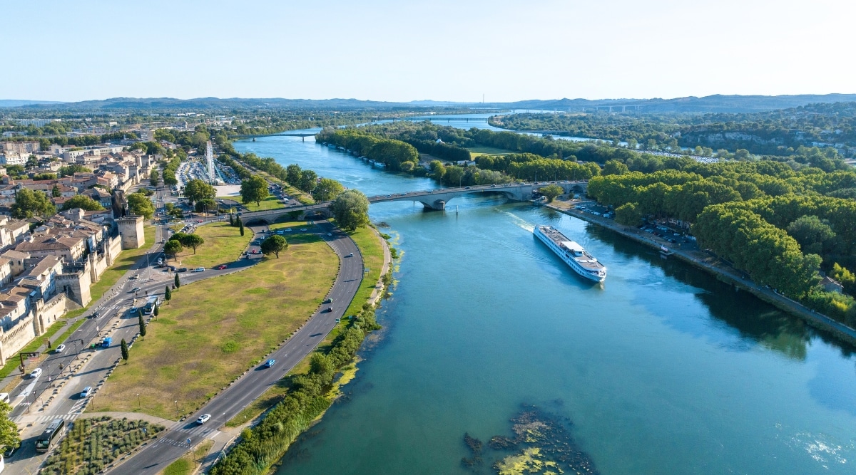 Aerial view of Ile de la Barthelasse. The Ile de la Barthelasse is a picturesque island in the Rhône River, near Avignon, France. This charming island is characterized by lush, rolling fields, vineyards, and countryside landscapes. A long, wide bridge connects the island with the city. A white cruise ship floats along the river.