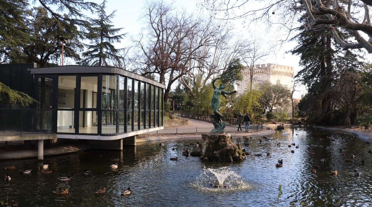 View of the Rocher des Doms park in Avignon, France. This peaceful oasis is characterized by lush green gardens, shaded walkways, and pristine laws. In the park there is a large Avignon Fountain which depicts a woman, described as a river nymph or a classical figure. She stands gracefully, holding a jar from which water flows, symbolizing the life-giving properties of water. Near the fountain there is a modern glazed building.