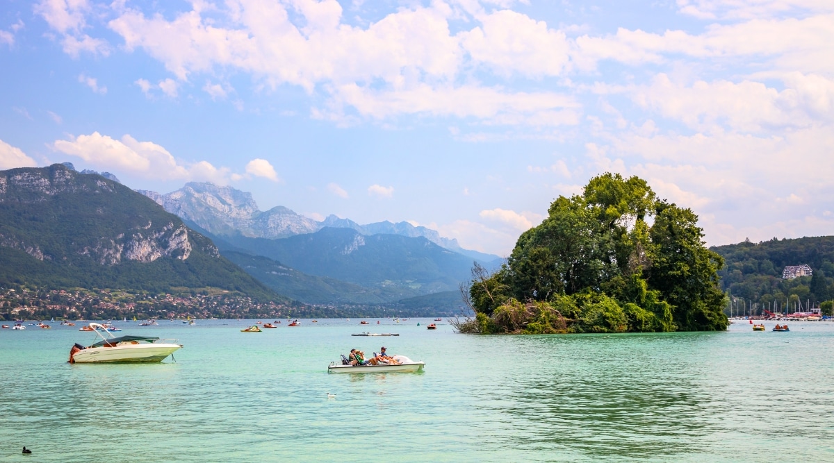 View of small wooded island in Lake Annecy. The small wooded island in Lake Annecy is a tranquil oasis of lush greenery, nestled amidst the glistening blue waters.The island's solitude is accentuated by the surrounding expanse of water. Many tourists on boats sail around it.