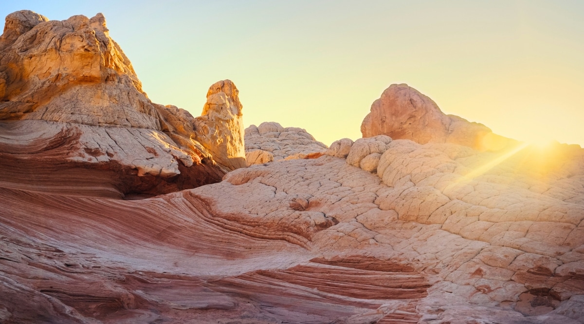 A image captures the enchanting beauty of a sunset over the Vermilion Cliffs. The sun, in its final moments before dipping below the horizon, bathes the landscape in a warm, golden glow. The cliffs, which are composed of intricate layers of red, orange, and white sedimentary rock, stand silhouetted against the radiant sky.