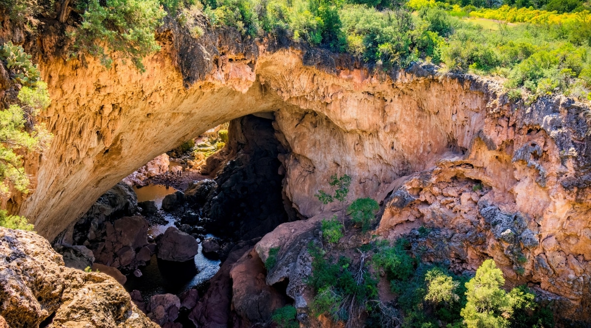 A captivating photo of Tonto Natural Bridge State Park. The image showcases the awe-inspiring natural bridge, a massive, weathered limestone formation, arching gracefully over a lush, green, and vibrant canyon. The surrounding lush vegetation and clear creek add to the park's natural charm.