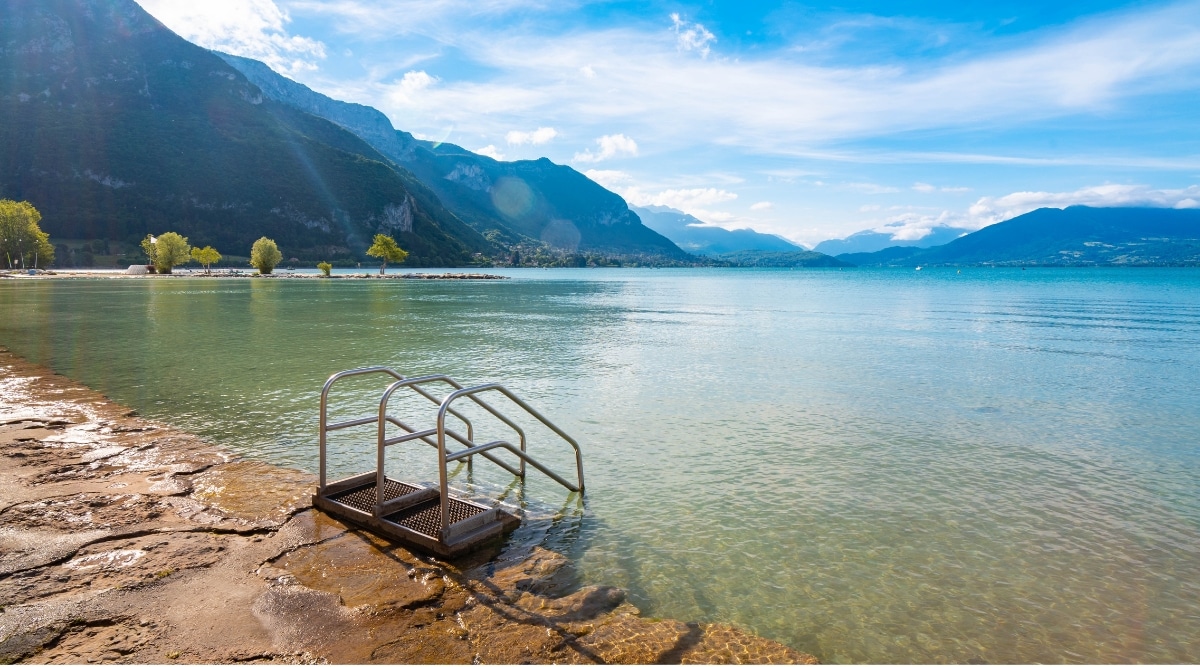 View of Annecy's beach in sunlight. Along the pristine shores of Lake Annecy, these sandy stretches are bordered by lush greenery and offer striking views of the surrounding French Alps. The water in the lake is crystal clear with shades of turquoise and emerald.