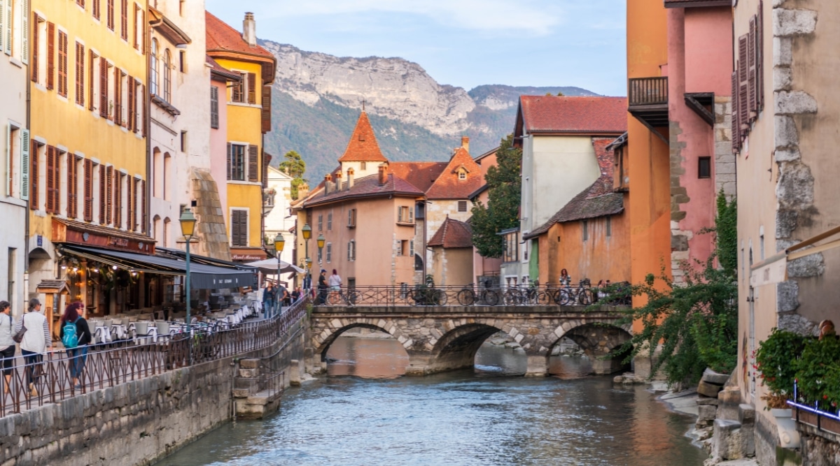 View of a street in the city of La Vieille Ville. Its narrow cobblestone streets wind through a maze of pastel-colored buildings while meandering canals lined with cafe terraces add to the town's romantic atmosphere. A small stone bridge in the form of three small arches connects the streets over the water canal.