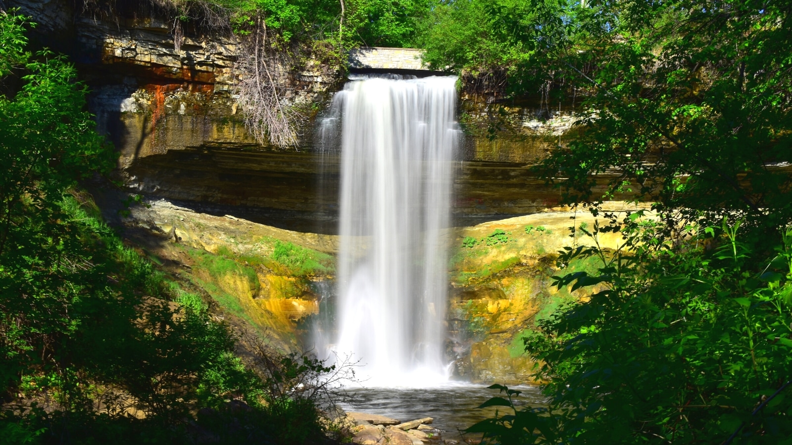 View of Minnehaha Falls, nestled within Minnehaha Park in Minneapolis. The waterfall cascades gracefully over a limestone ledge, creating a mesmerizing drop into a clear pool below. Surrounded by lush greenery, the falls are embraced by a picturesque setting of towering trees.