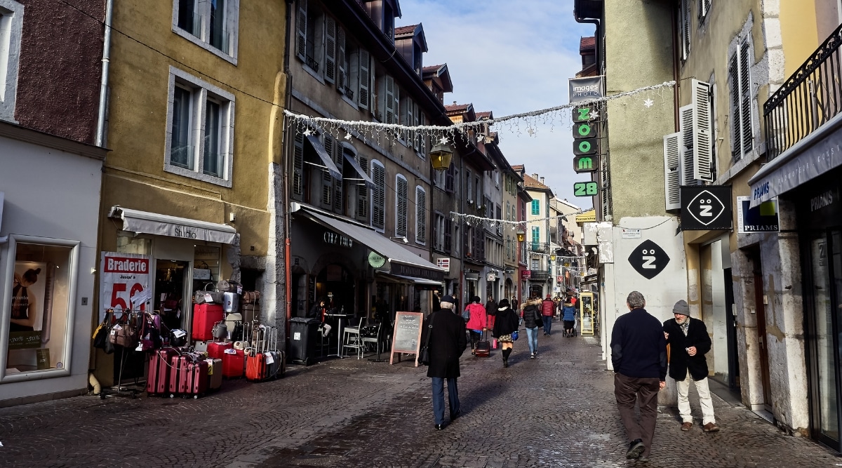 View of one of the streets in Annecy France. The street is narrow, with old colorful houses, various shops and restaurants along it. People walk along the street.