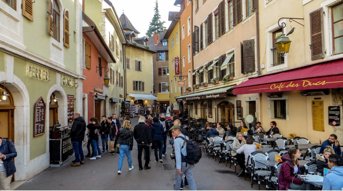 One of the narrow and crowded streets in Annecy France. The houses have 3-4 floors, with wooden shutters on the windows. On the lower floors there are various restaurant terraces where people relax.