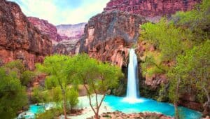 An awe-inspiring view of Havasu Falls. The image reveals the stunning turquoise-blue waters plunging over the red-hued cliffs into a crystal-clear pool below. Surrounded by lush greenery, the waterfall creates a breathtaking contrast against the rugged desert landscape.