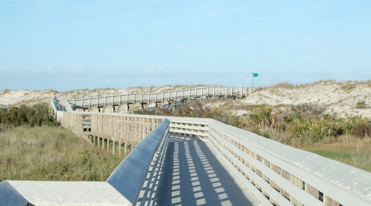 The photograph depicts the approach to the beach via a lengthy white wooden bridge in Anastasia State Park. The bridge extends over the landscape, providing access to the sandy shores. Surrounding vegetation and natural elements frame the path to the beach.