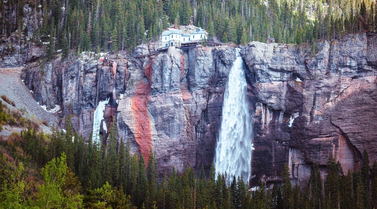 A photograph capturing Bridal Veil Falls in Colorado. The image showcases the waterfall cascading down a rocky cliff, surrounded by natural vegetation. The rocky terrain and forested backdrop contribute to the scenic landscape. 
