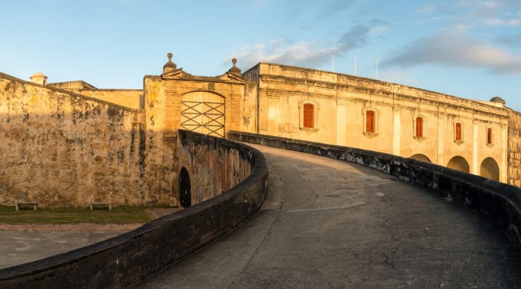 A photograph capturing the entrance of Castillo San Cristobal. The image features the fortified structure's main gate and entrance pathway. Stone walls and architectural elements are visible. The entrance is adorned with classic design elements.