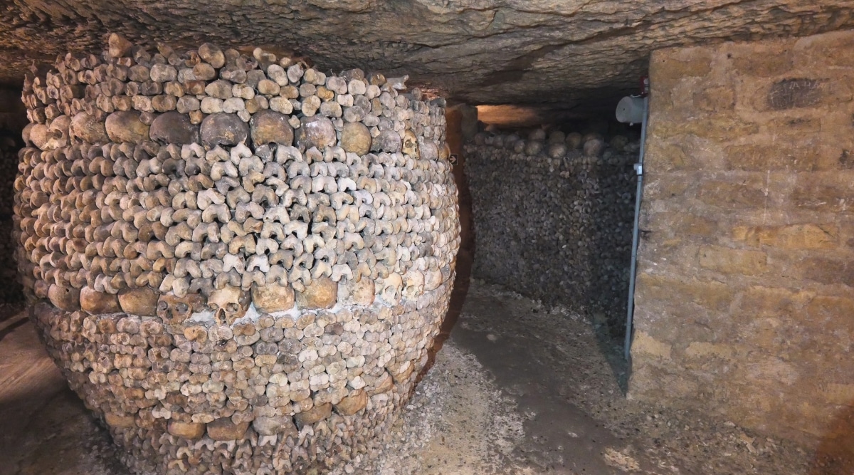 Catacombs in Paris. The image showcases the dimly lit, underground passages adorned with neatly stacked human skulls and bones. The meticulous arrangement of bones forms intricate patterns.