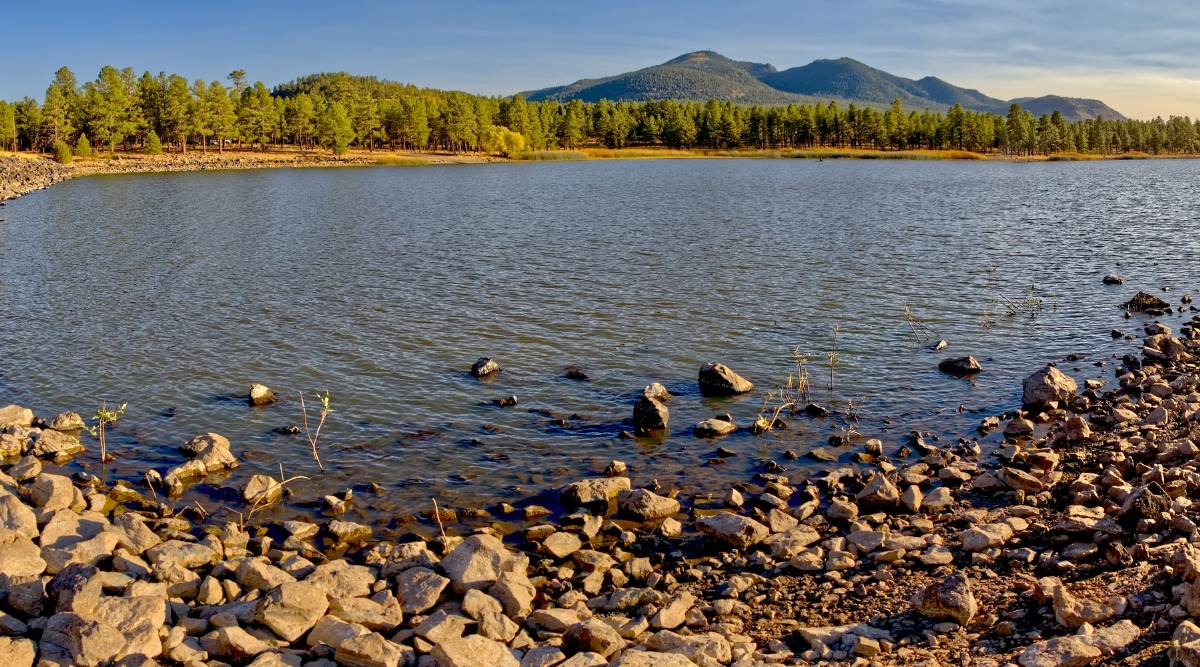A picturesque photograph showcases Cataract Lake in Arizona. The expansive lake boasts a wide expanse of fine gravel along its shore, with numerous trees dotting the landscape in the distance.