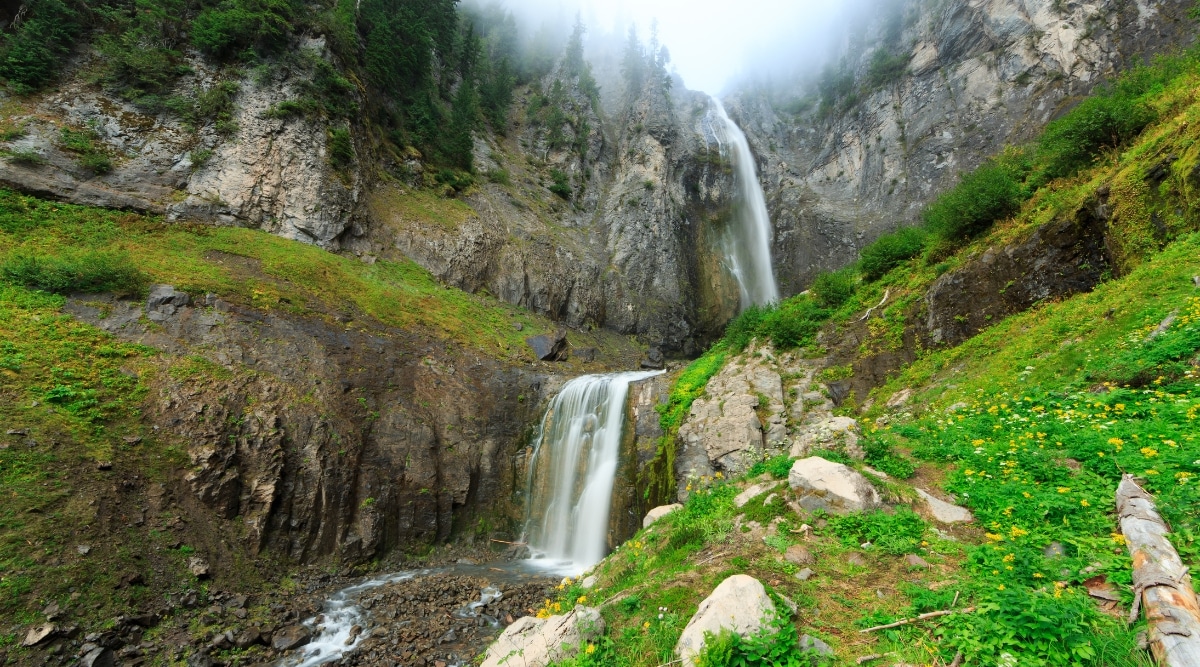 A photograph illustrating Comet Falls in Washington. The image captures the waterfall cascading down a rocky cliff surrounded by lush greenery. The falls present a picturesque scene with water flowing into a pool below. The landscape features a mix of trees and rocky formations.
