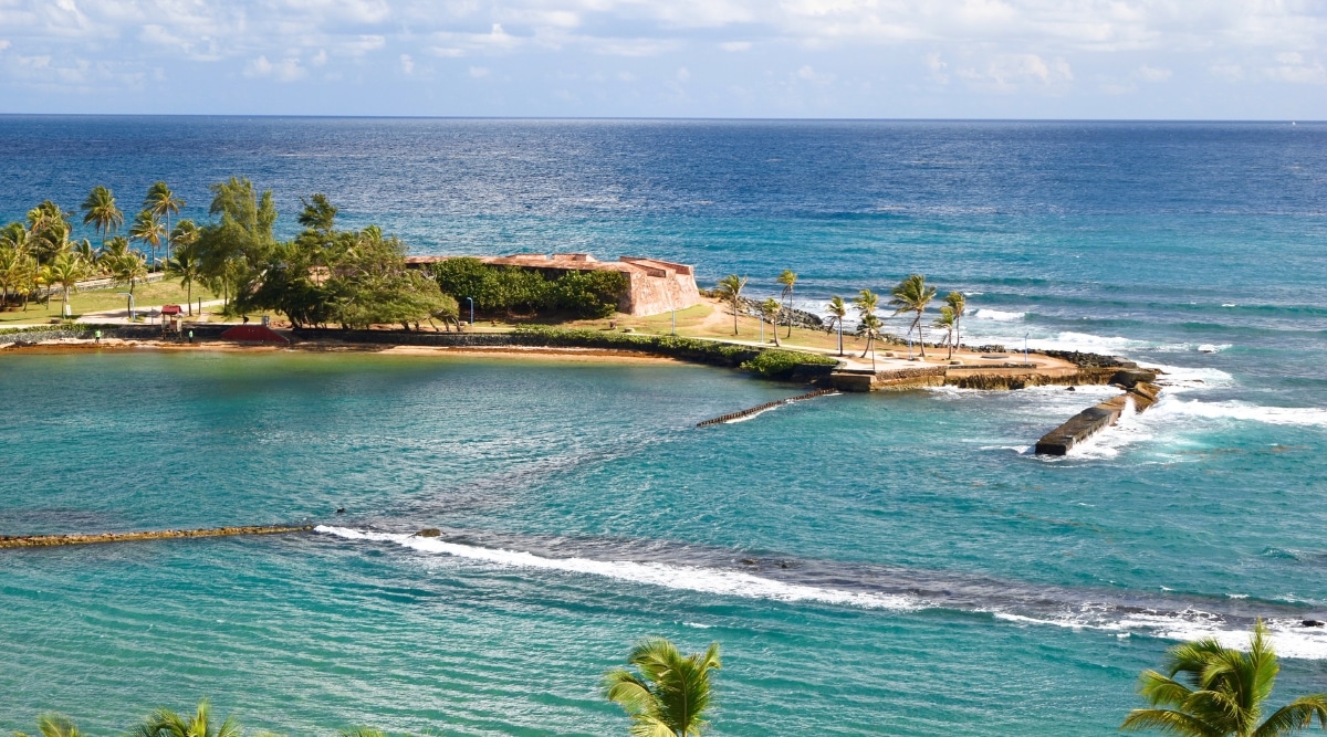 A photograph illustrating Escambron Beach in Puerto Rico. The image captures the beach's shoreline and sandy expanse, bordered by the clear waters of the Atlantic Ocean. The surrounding area features palm trees and coastal vegetation.