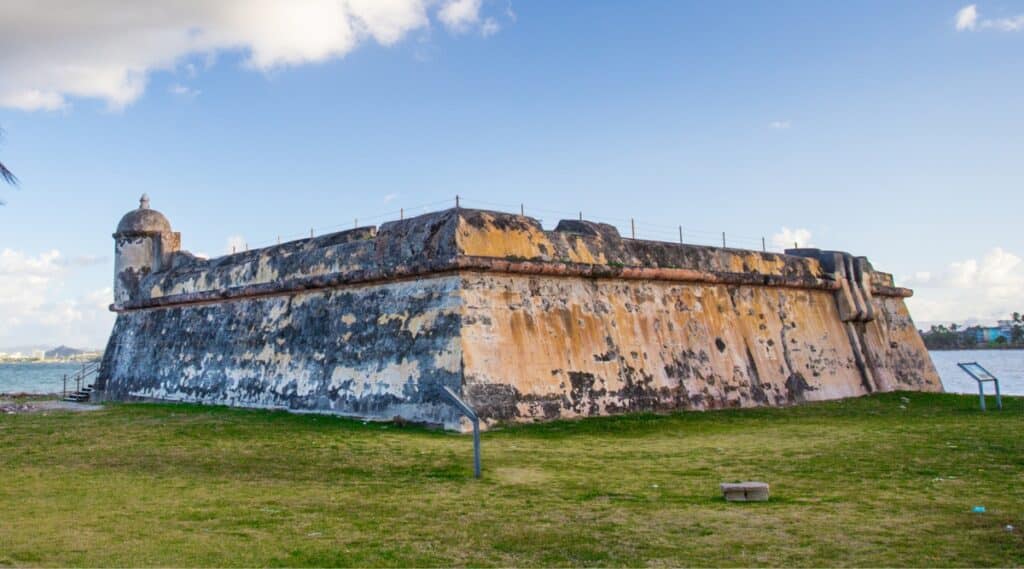 A photograph capturing Fortín San Juan de la Cruz in Puerto Rico. The image features the historic fortress situated against a coastal backdrop. The fort exhibits traditional military architecture with solid stone walls and structures.