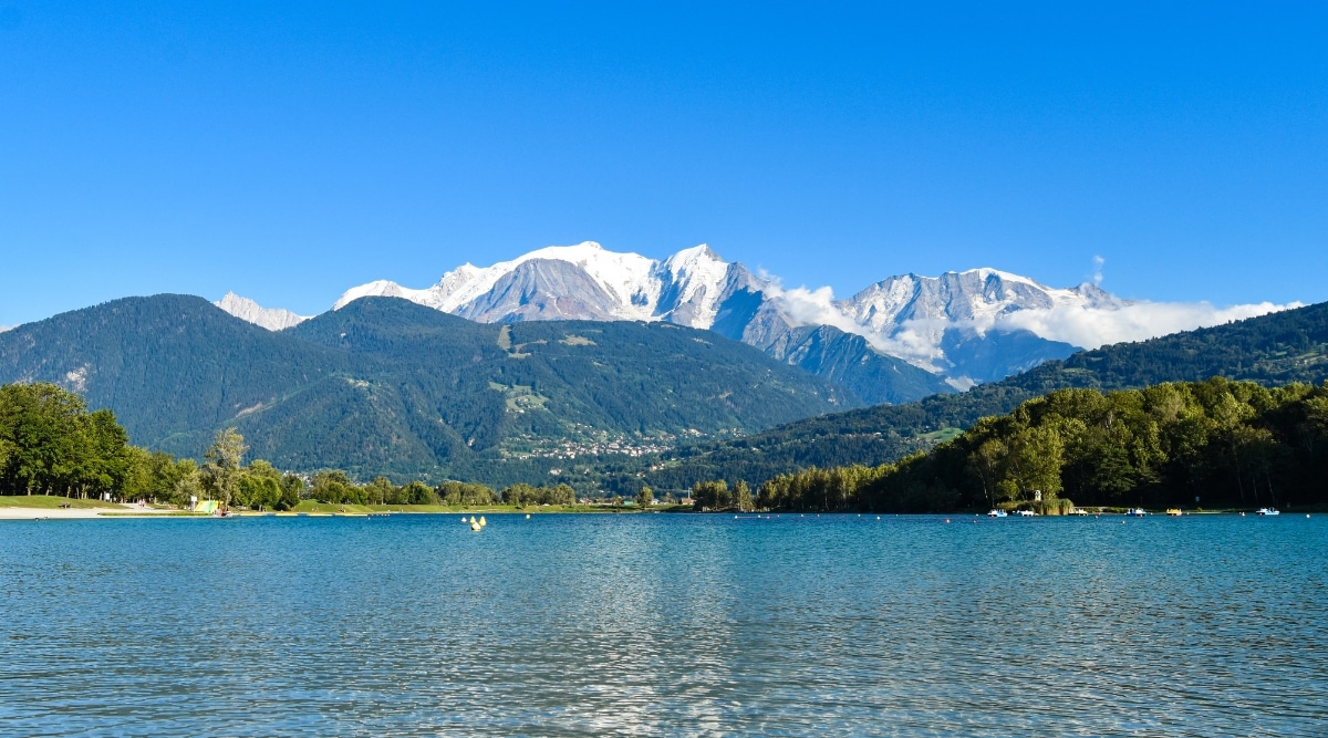 Lac de Passy in the Chamonix region of France. The crystal-clear waters of the lake reflect the surrounding alpine landscape, mirroring the towering peaks, dense forests, and azure sky above. The distant mountain range frames the horizon.