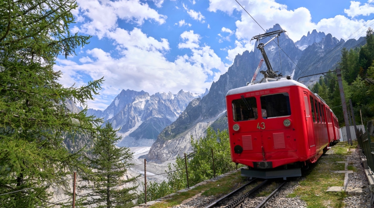 An evocative image captures the historic Montenvers Railway in Chamonix, France. The iconic cogwheel train ascends a steep mountainside. The railway, surrounded by towering peaks and dense evergreen forests. As the train winds its way upward, passengers are treated to panoramic views of snow-capped mountains, alpine meadows.