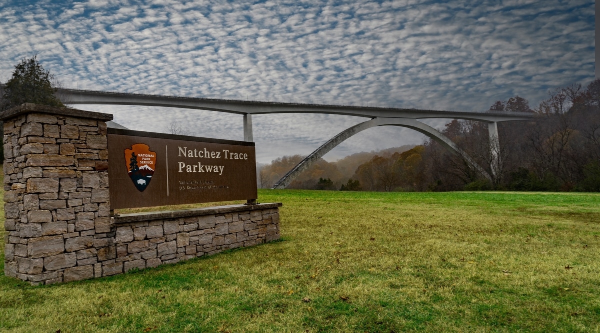 Close up of the Natchez Trace Parkway sign on a cloudy day with an arch bridge in the background in Tennessee. 