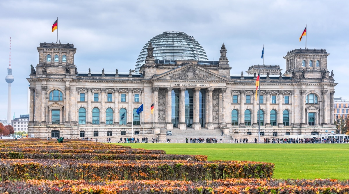 A striking photograph capturing the iconic Reichstag building in Berlin. The image showcases the historic architecture of the Reichstag, featuring its grand dome and neoclassical facade against a clear sky. The German national flag flutters atop the dome.