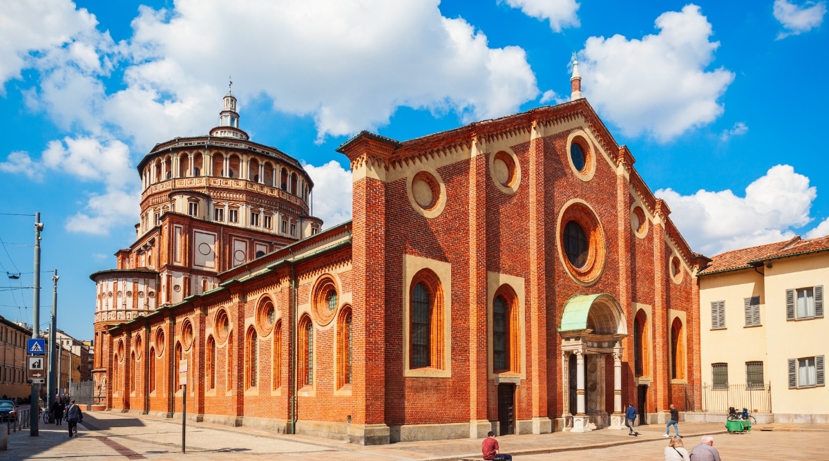 A photograph capturing the architectural splendor of Santa Maria delle Grazie in Milan. The image showcases the exquisite facade of the church, a harmonious blend of Gothic and Renaissance styles, adorned with intricate sculptures and detailing. The warm hues of the building's exterior contrast beautifully with the blue sky overhead.