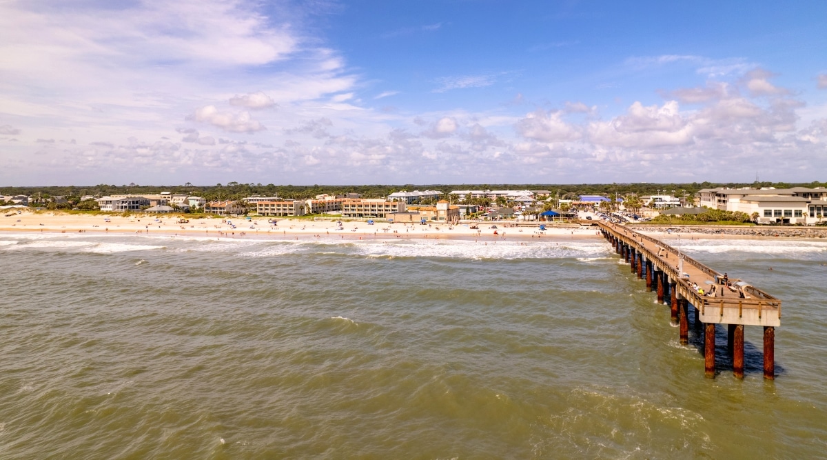 Aerial view from a quadcopter capturing the ocean side beach in St. Augustine, Florida, featuring a long pier extending over the coastline. The image features the coastal landscape with sandy shores. Beachgoers and umbrellas are visible along the shoreline. The coastal cityscape provides a backdrop.