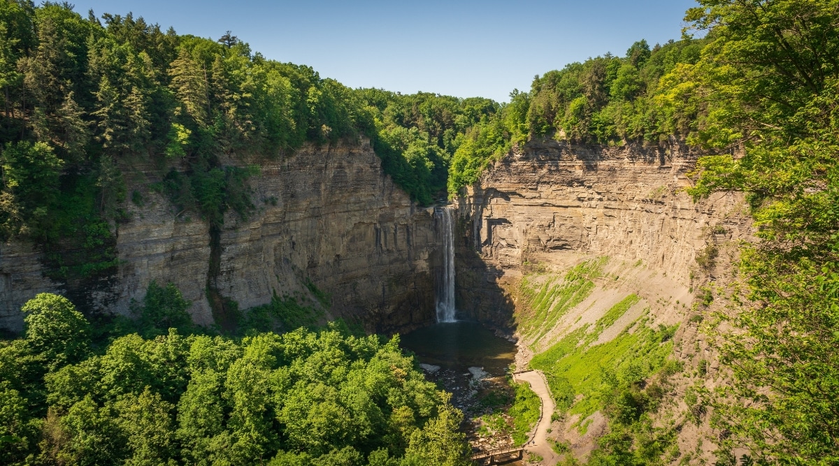 A photograph capturing Taughannock Falls in New York. The image features the waterfall cascading down the rock face, framed by surrounding trees and rocky formations. The landscape is characterized by lush greenery and the natural contours of the rock formations.