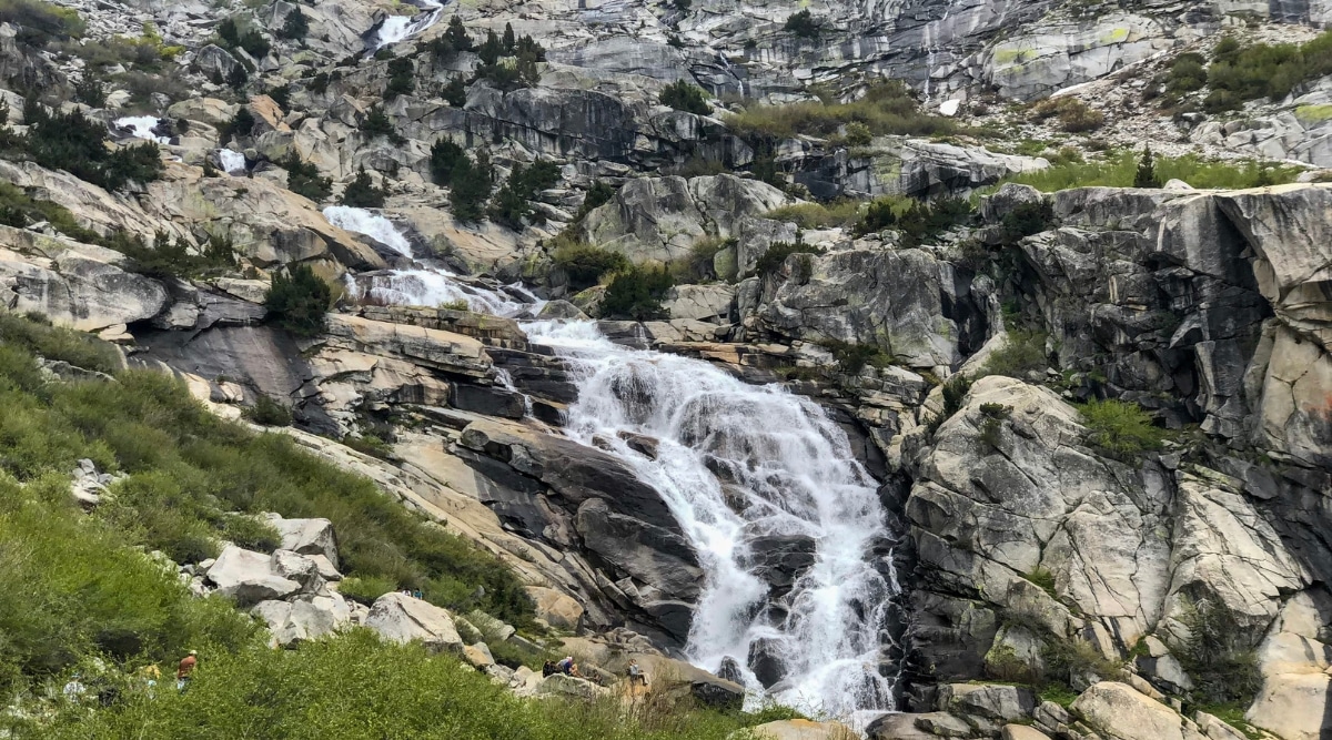 A photograph illustrating Tokopah Falls in California. The image portrays the waterfall cascading down a rocky landscape surrounded by dense vegetation. The surrounding terrain consists of rugged rocks and greenery.