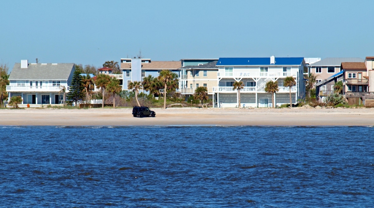 A photograph of Vilano Beach in St. Augustine, Florida, reveals the sandy coastline set against the backdrop of expansive beachfront houses. A black SUV can be seen driving along the sandy shore.