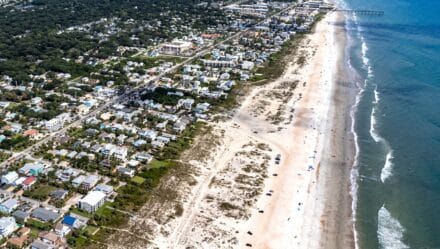 The aerial photograph captures the panoramic view of St. Augustine Beach in Florida. The image presents a serene coastal landscape, with the sandy shore extending seamlessly towards the horizon. The sandy expanse is dotted with beachgoers and umbrellas.