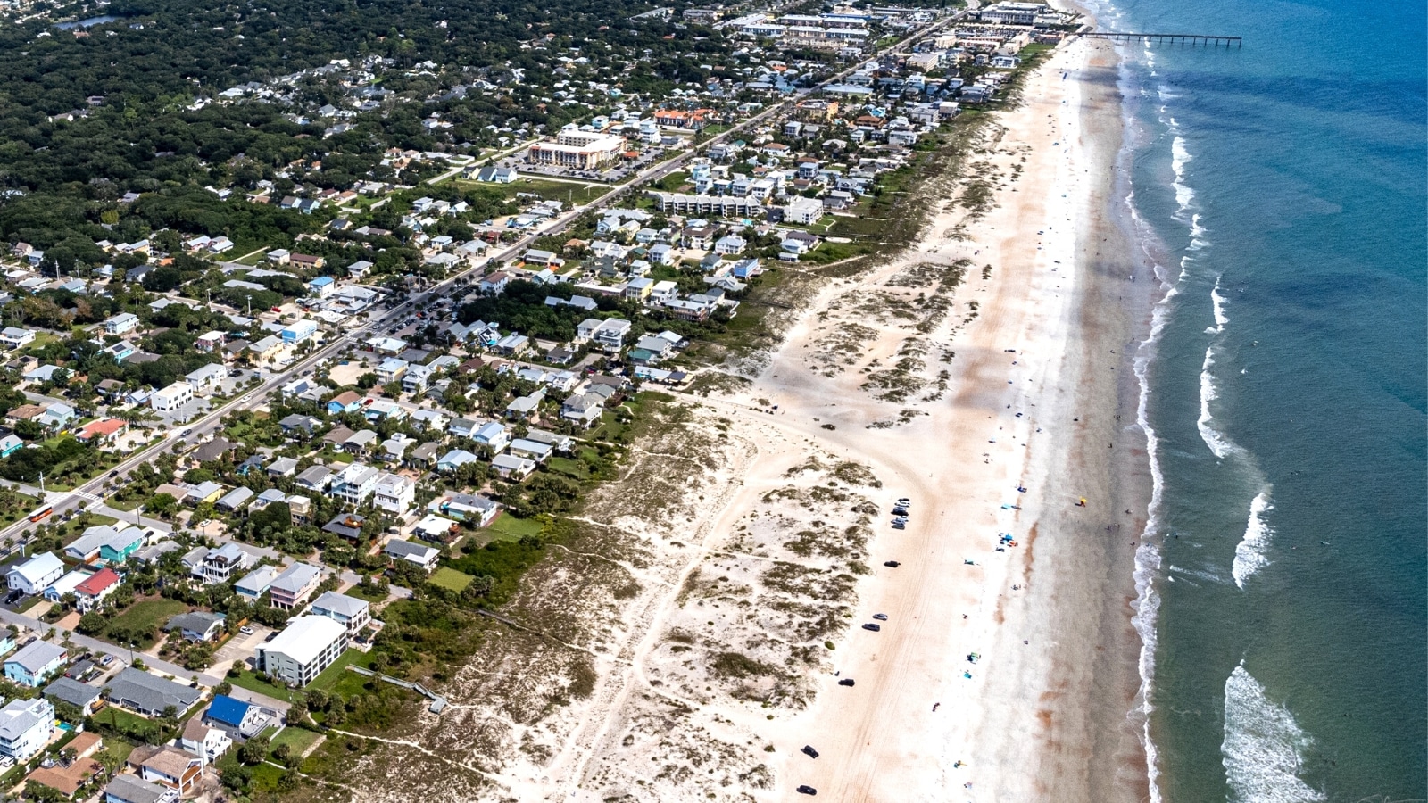 The aerial photograph captures the panoramic view of St. Augustine Beach in Florida. The image presents a serene coastal landscape, with the sandy shore extending seamlessly towards the horizon. The sandy expanse is dotted with beachgoers and umbrellas.