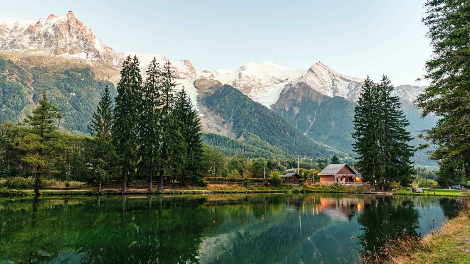 A breathtaking panorama of majestic Alps peaks, snow-capped summits, and pristine landscapes. View of lakes with clear water reflecting tall green pine trees, the Alps and two small huts.