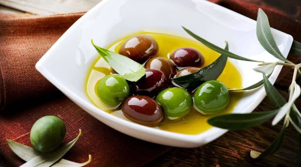 A photograph displaying plates containing green and red olives immersed in oil. The olives are presented against a neutral background, showcasing their colors and textures.