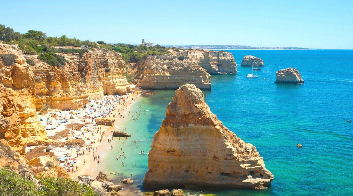 A photograph capturing the landscape of Algarve, Portugal. The image features a coastal scene with notable geographical features such as cliffs and beaches. The coastal terrain is marked by its characteristic topography, and the sea is visible in the background. There are many visitors on the beach with beach umbrellas.