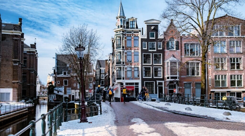 A photograph capturing a winter scene on a street in Amsterdam, Netherlands. The image depicts snow-covered surroundings with individuals engaging in various activities. The buildings lining the street are characterized by their typical three to four-story architectural structure.Pedestrians can be observed navigating the snow-covered paths.