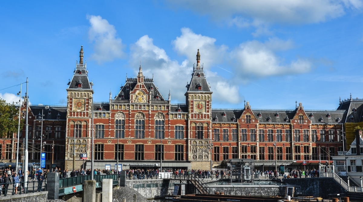 A depiction of Centraal Station in Amsterdam, a prominent transportation hub. The image captures the exterior of the station, showcasing its architectural features, including a combination of classic and modern elements. Commuters and pedestrians can be observed in the vicinity.