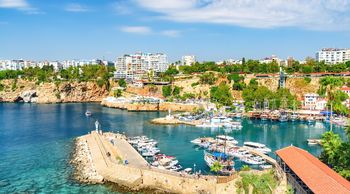 A photograph showcasing the city of Antalya, Türkiye. The image features a cityscape with a prominent marina, where ships are anchored. 