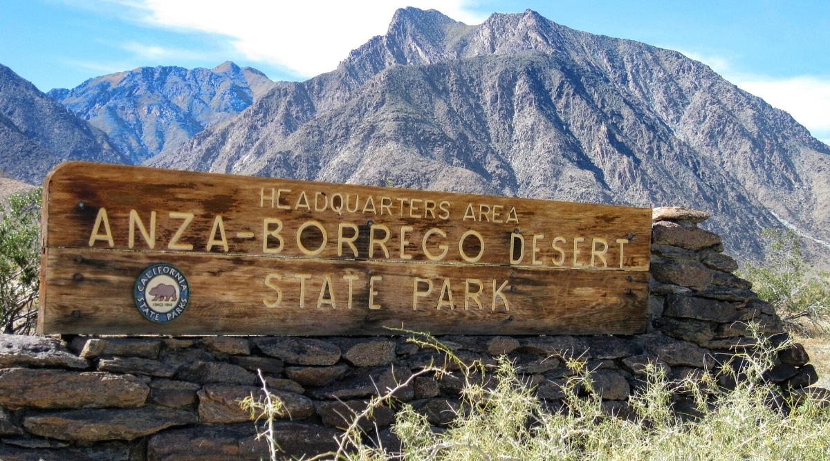 A photograph depicting a wooden stele with the inscription 'Anza-Borrego Desert State Park' affixed to stonework. The image is situated against the backdrop of a mountain. The stele serves as a marker, providing information about the location and denoting the boundary of Anza-Borrego Desert State Park.
