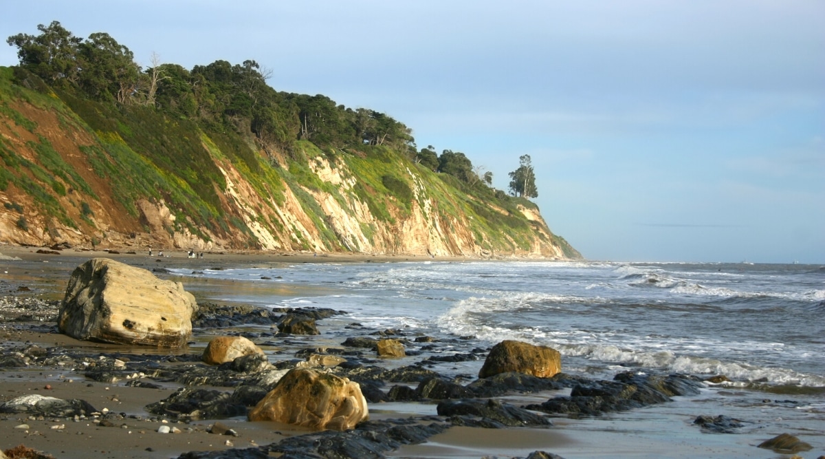 A view of Arroyo Burro Beach in Santa Barbara featuring the ocean with sizable waves. To the left of the frame is a lengthy hill adorned with greenery, and on the beach, cobblestones are visible. The image provides a straightforward depiction of the coastal landscape, showcasing the natural elements of the beach, including the ocean, hill, and stony shore.