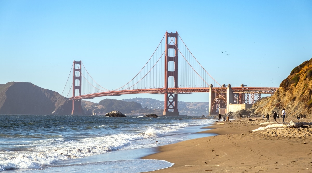 A direct view of Baker Beach in San Francisco, featuring a shoreline on the right where individuals can be seen walking along the beach. In the distance, a prominent bridge is directly visible.