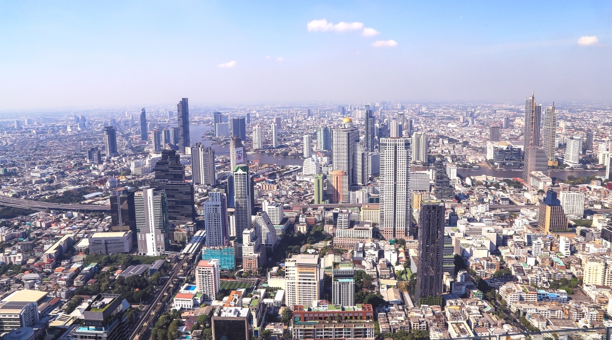 A drone view capturing the cityscape of Bangkok during daylight. The image showcases numerous high-rise buildings with varying floor counts, contributing to the city's skyline. The presence of several clouds in the sky indicates typical atmospheric conditions.