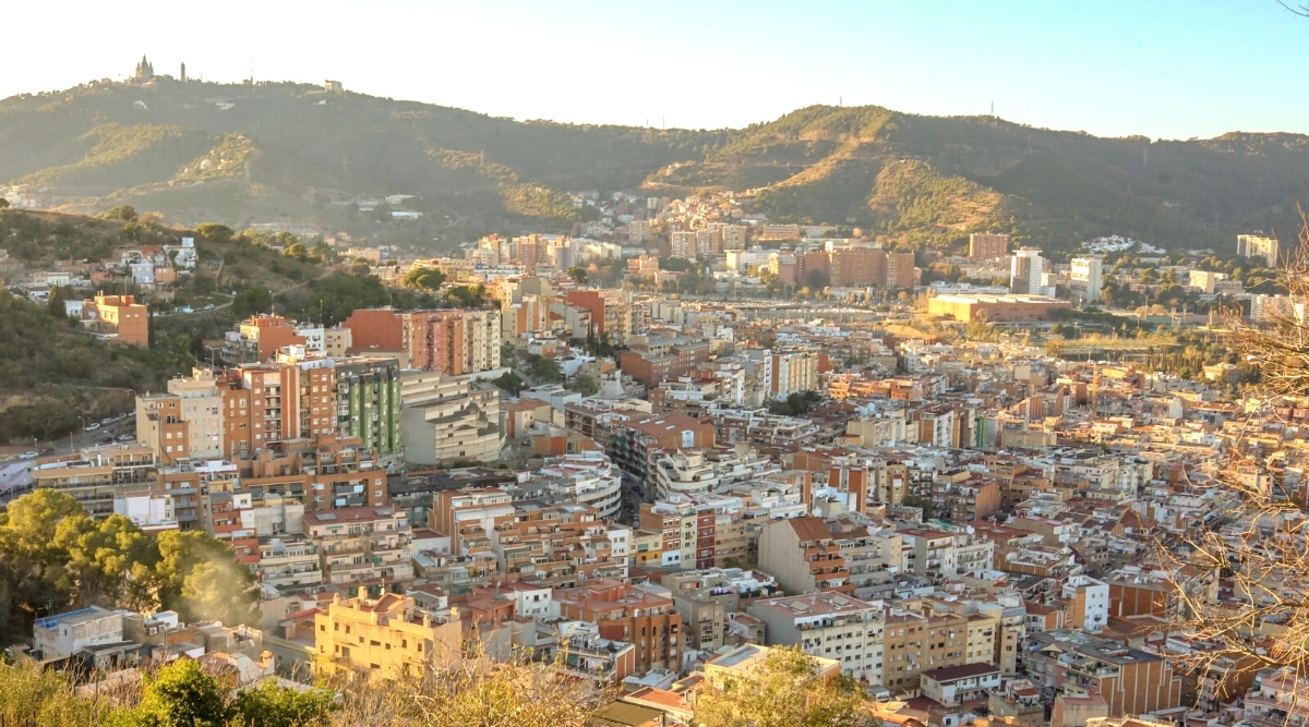 An aerial photograph capturing the cityscape of Barcelona, Spain. The image showcases the urban layout, featuring an array of buildings, streets, and public spaces. The weather conditions in the image appear clear, with minimal cloud cover.