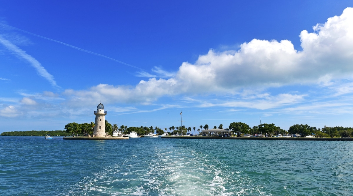 A direct view capturing Biscayne National Park in Florida. The image showcases a prominent lighthouse against a backdrop of white clouds in the sky. The park features abundant greenery, and the expansive ocean is visible in the foreground. 