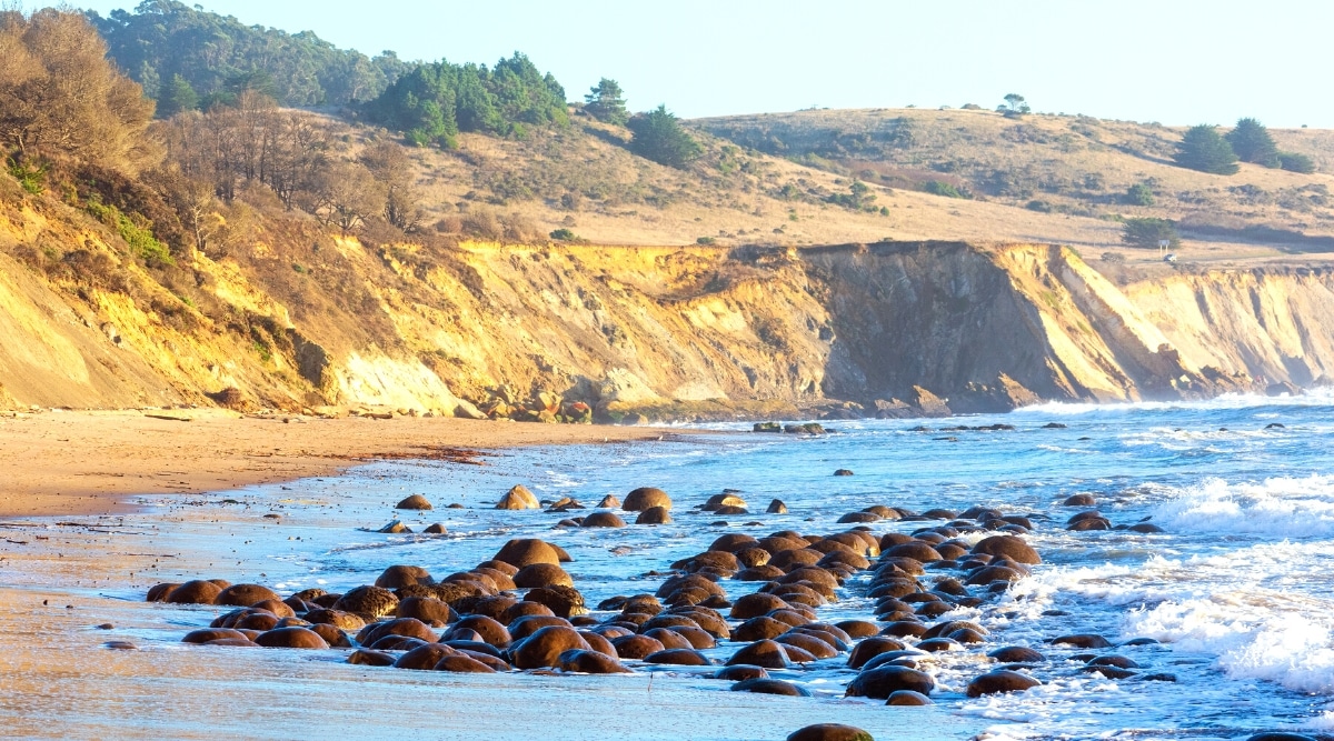 The photograph captures the scene at Bowling Ball Beach in Mendocino. In the foreground, small boulders are scattered along the shore, partially submerged in the water. The background features sandy rocks, creating a textured coastal landscape.