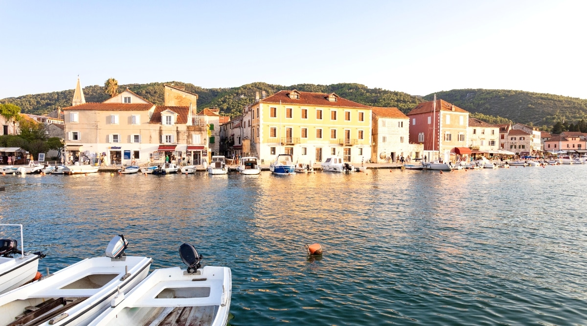A view capturing the coastal landscape of Brac, Croatia. The image features a cluster of three- to four-story buildings in close proximity to the sea, with numerous white boats visible along the shoreline. Hills provide a backdrop to the scene.