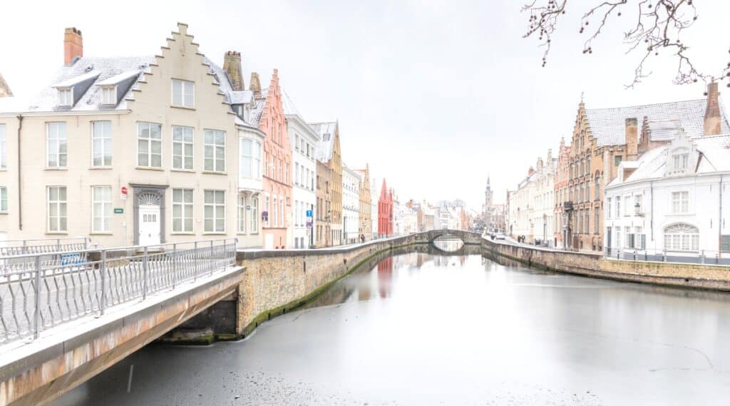 A photograph depicting a cold winter day in Bruges, Belgium, with light snowfall. The image captures a water canal stretching along a city street, devoid of people. The architecture features houses of various colors, including orange, white, beige, and red. 