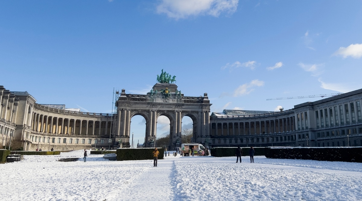 A view of Cinquantenaire Park covered with snow in Brussels. The image captures the park's landscape blanketed in snow, and few people are visible in the scene. The sky is clear with white clouds.