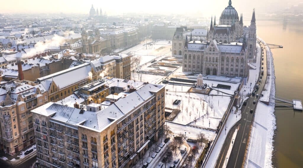 An aerial view of the snowy riverside of Pest, featuring the prominent landmarks of the Parliament of Hungary and St. Stephen's Basilica. The image captures the misty ambiance of a winter morning in Budapest. The snow-covered landscape adds a seasonal touch to the cityscape.The Parliament of Hungary stands out with its distinctive architecture, while St. Stephen's Basilica contributes to the overall skyline. 