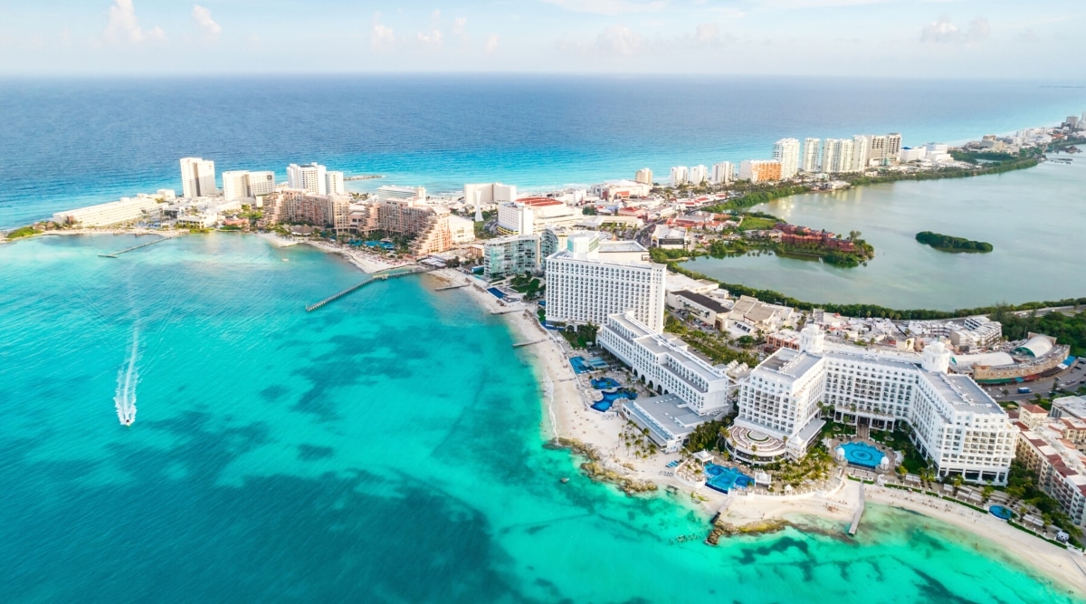 An aerial photograph capturing Cancun, Mexico, from a top-down perspective. The image showcases the city's urban layout, including roads, buildings, and coastal features. The recognizable hotel zone is visible, characterized by a concentration of resorts along the shoreline. The weather appears clear, offering a straightforward depiction of the cityscape.